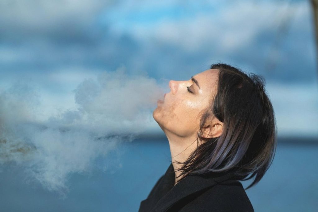 Young woman exhaling vapour outdoors against a blue sky, illustrating vape use in the UAE